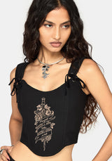 A woman wearing a black sleeveless corset with side tie details and a beige graphic print on the front, photographed against a light gray studio background.