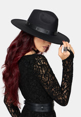 A woman with long red hair wearing a black wide-brim fedora hat with a textured band, a black lace long-sleeve top, and a black belt, shown against a light gray background.