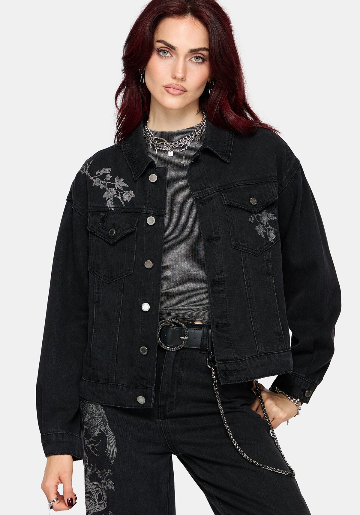 A model with dark red hair wearing a black denim jacket with gray floral embroidery on the chest, layered over a gray textured top and black jeans.
