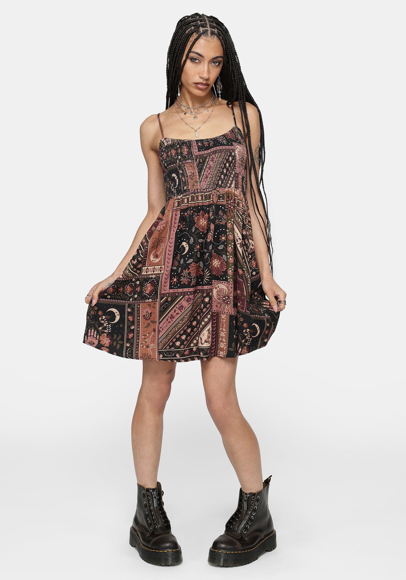 A woman modeling a sleeveless spaghetti-strap mini dress with a bohemian-style patchwork print in black, rust, and dusty pink, paired with black combat boots against a light gray studio background.