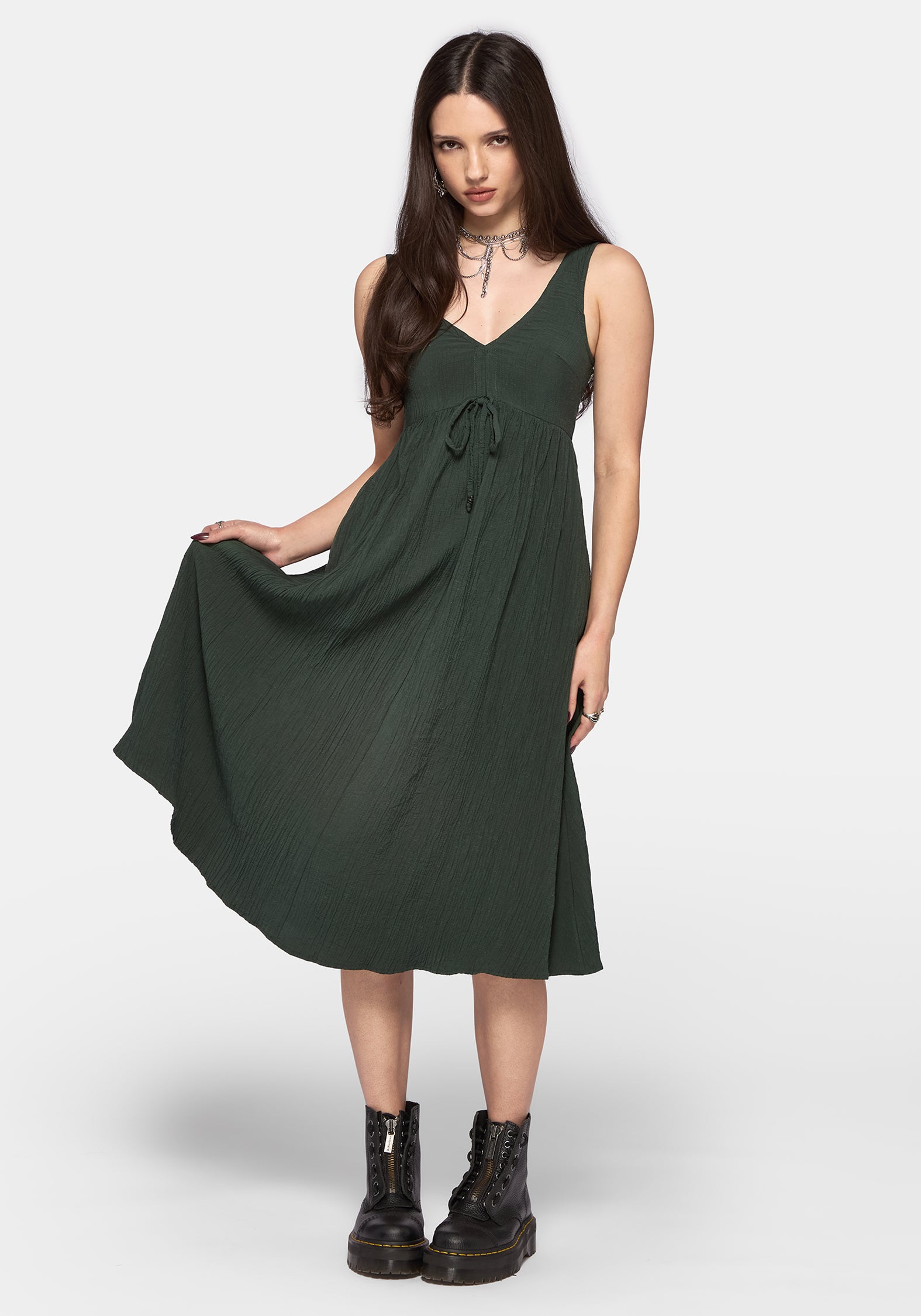 A woman modeling a dark forest green sleeveless midi dress with a V-neckline and drawstring waist, paired with black combat boots against a light gray studio background.