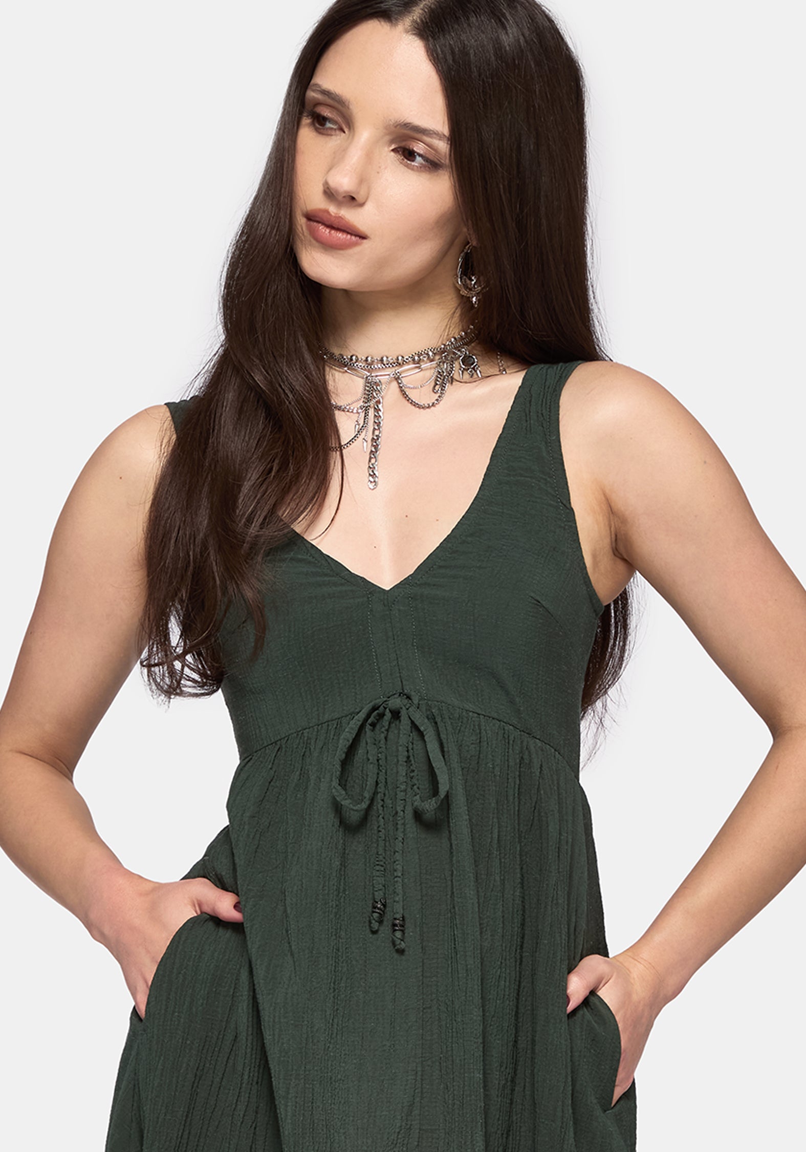 A woman modeling a sleeveless dark green V-neck dress with a drawstring waist and side pockets, set against a plain light gray background.