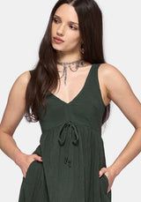 A woman modeling a sleeveless dark green V-neck dress with a drawstring waist and side pockets, set against a plain light gray background.