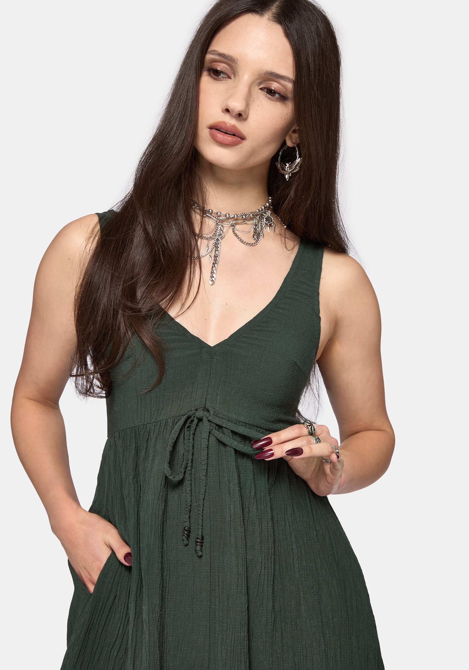 A woman modeling a sleeveless dark green V-neck dress with a tie waist and pleated skirt, set against a plain light gray background.