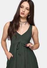 A woman modeling a sleeveless dark green V-neck dress with a tie waist and pleated skirt, set against a plain light gray background.