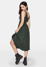 A woman modeling a dark green sleeveless midi dress with a gathered waist and side slit, paired with black ankle boots, against a light gray studio background.