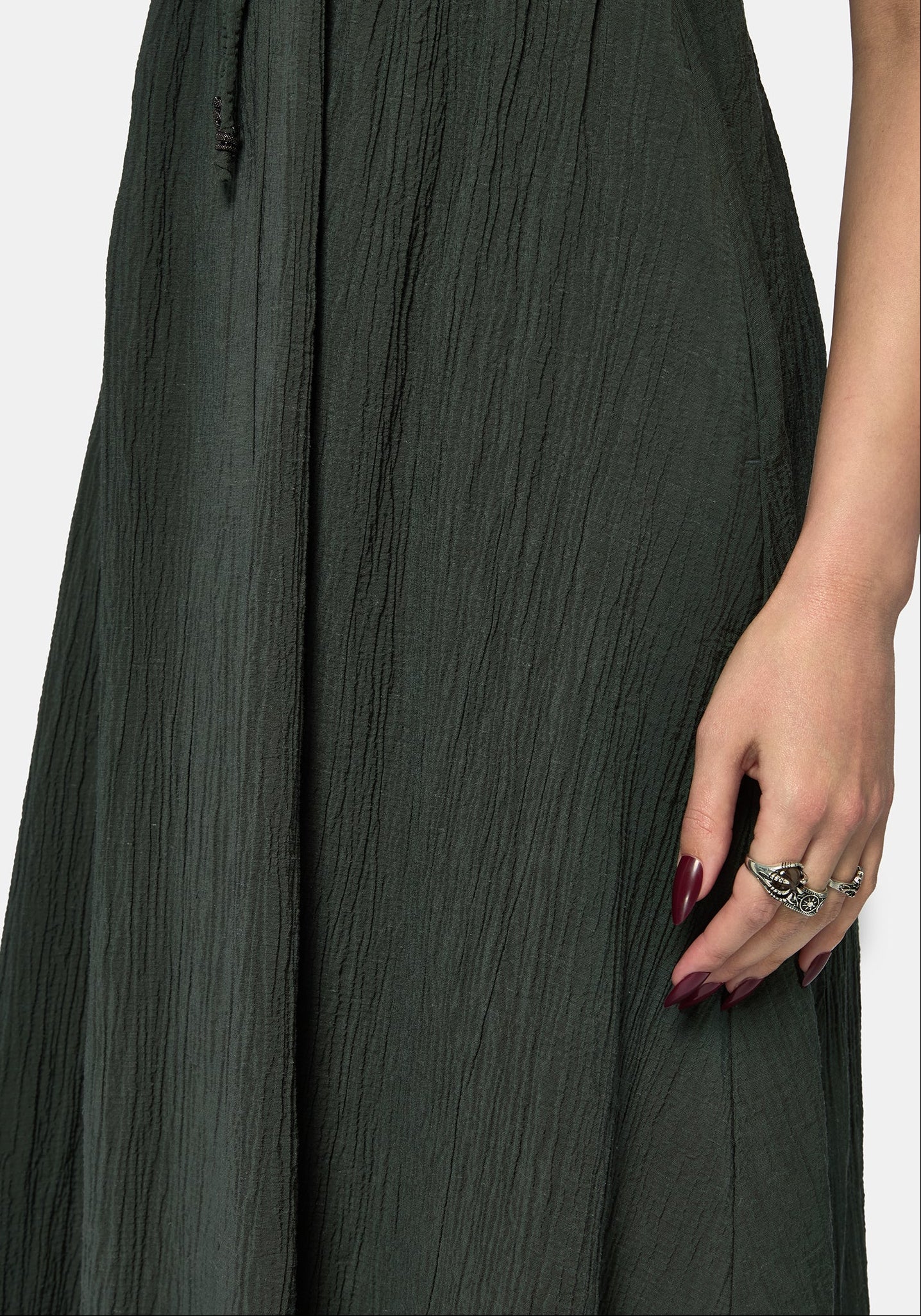 Close-up of a dark olive green crinkled maxi dress with a drawstring waist, worn by a model whose hand and manicured nails are visible on the right side.