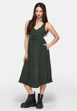 A woman modeling a sleeveless dark green V-neck dress with a tie waist and pleated skirt, set against a plain light gray background.