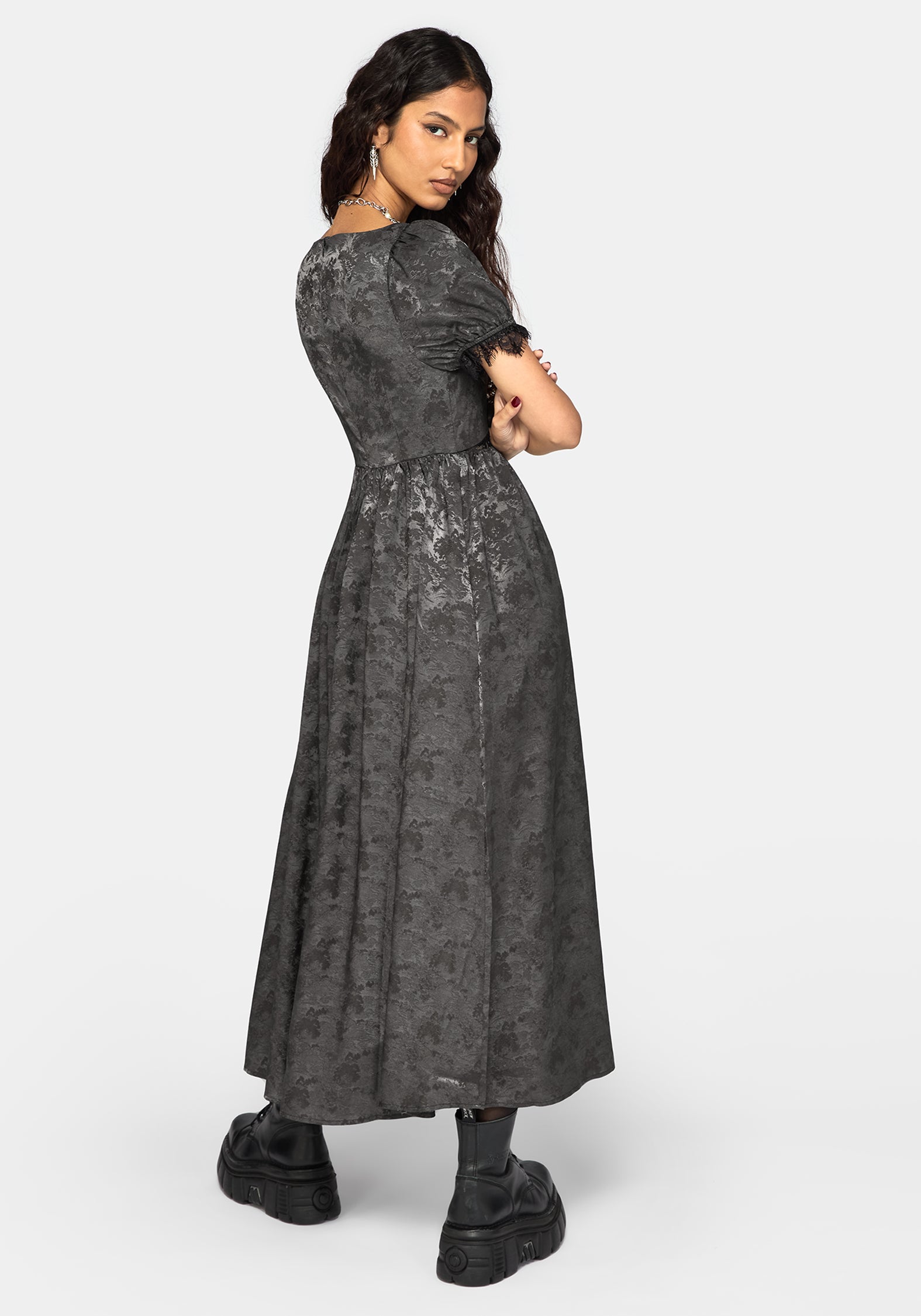 A woman modeling a dark gray short-sleeved midi dress with a textured, lace-like pattern, paired with black boots, against a light gray studio background.