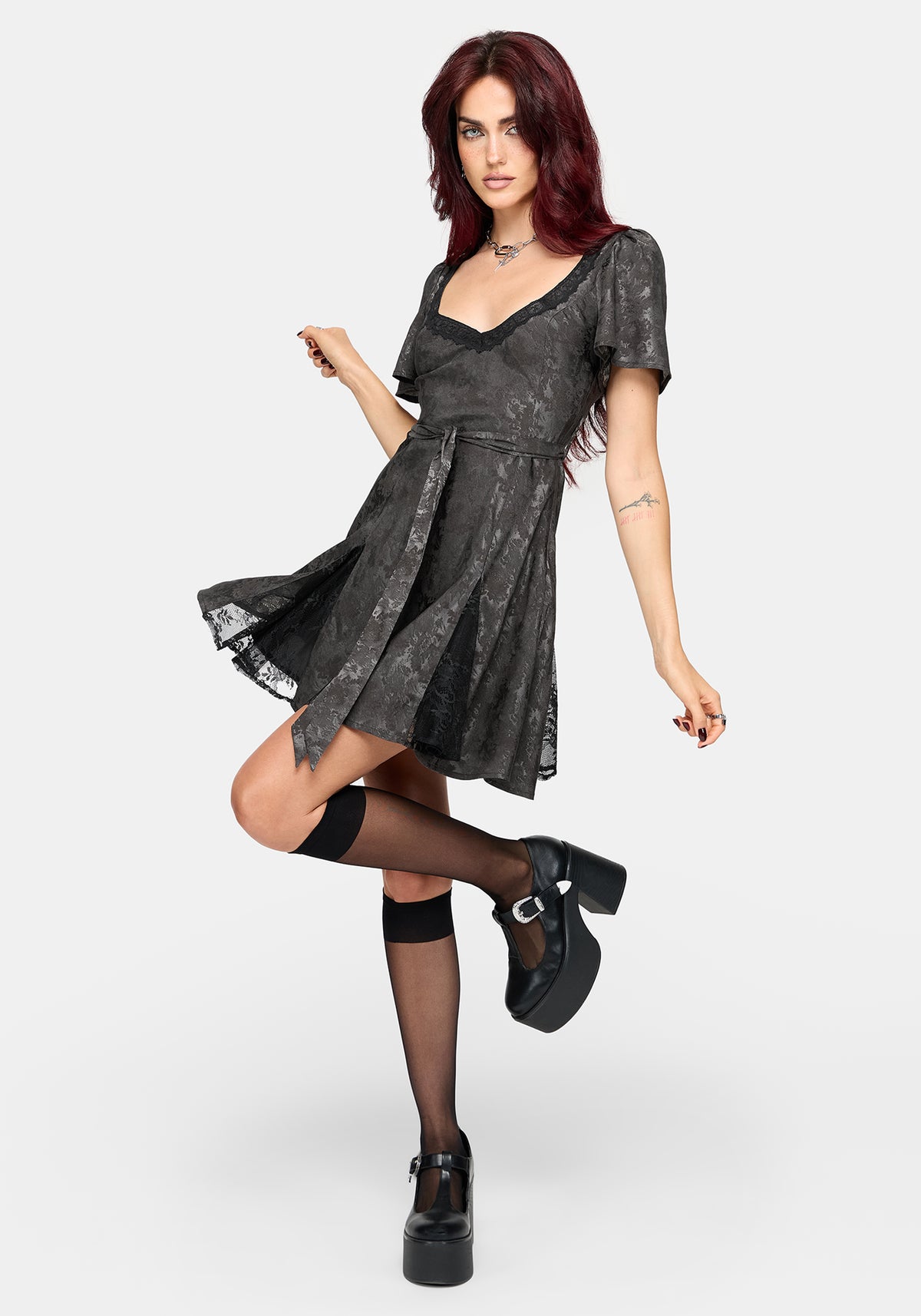 A model with red hair wearing a short-sleeved grey mini dress, black thigh-high stockings, and black platform Mary Jane shoes, posing against a light gray studio background.