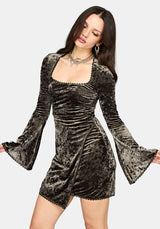 A woman modeling a short green crushed velvet mini dress with long bell sleeves, a square neckline, and a ruched bodice against a light gray background.