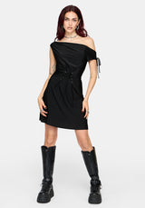 A woman modeling a black one-shoulder mini dress with a side tie detail, holding a black studded shoulder bag against a light gray background.