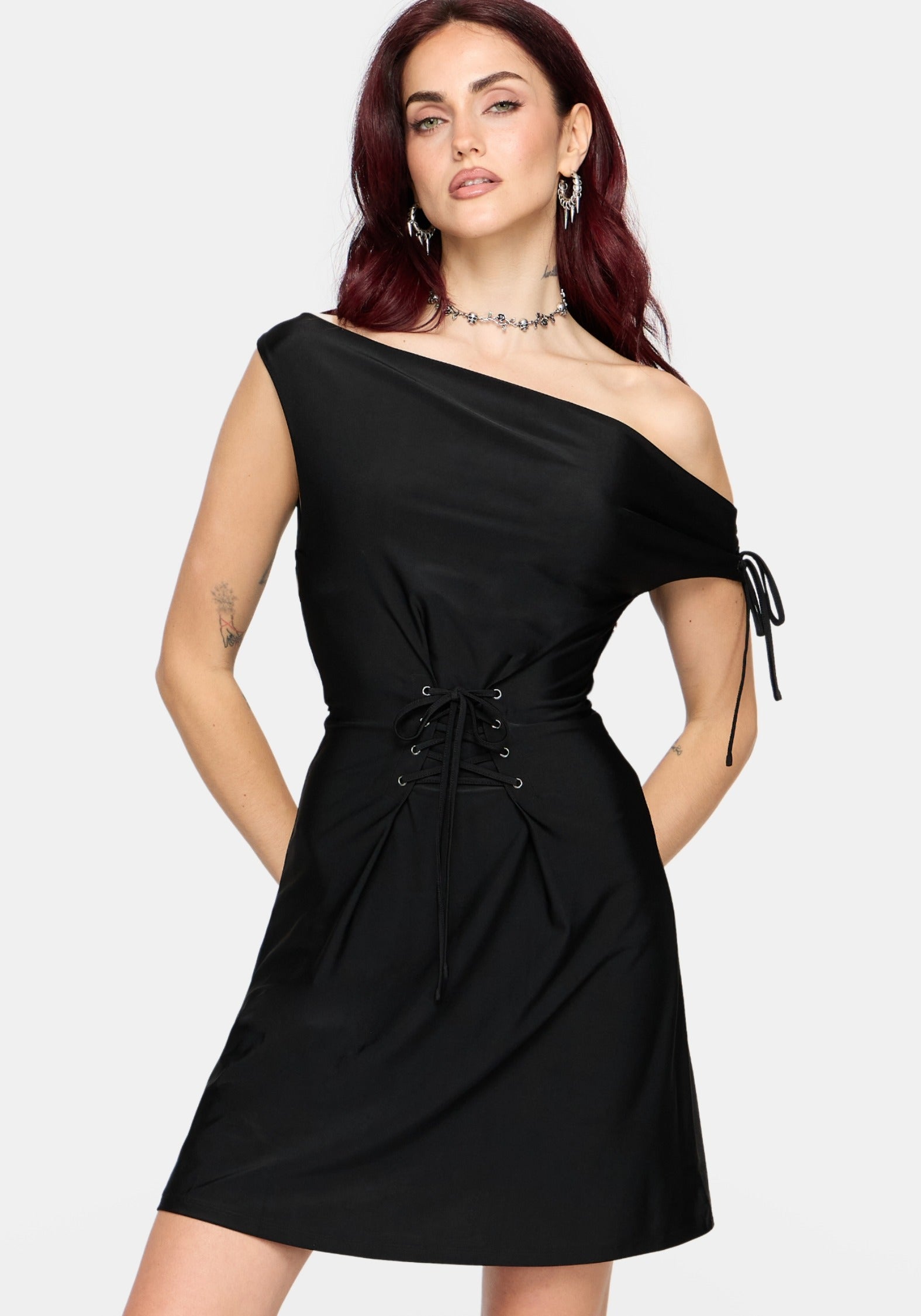 A woman modeling a black one-shoulder mini dress with a side tie detail, holding a black studded shoulder bag against a light gray background.