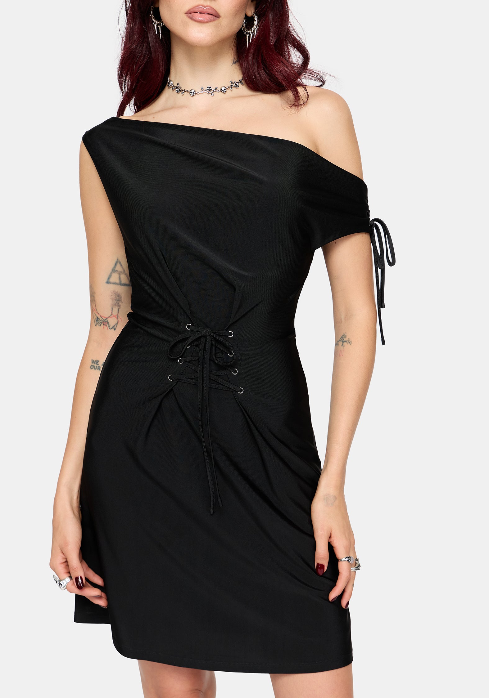 A woman modeling a black one-shoulder mini dress with a side tie detail, holding a black studded shoulder bag against a light gray background.