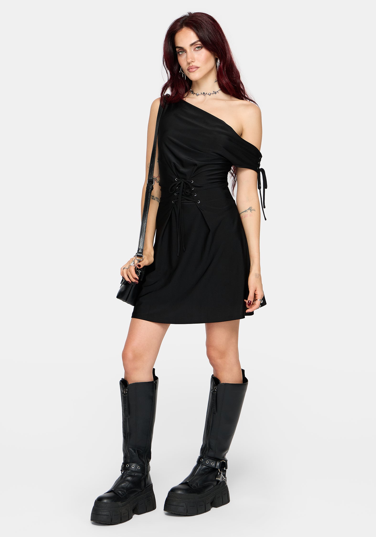 A woman modeling a black one-shoulder mini dress with a side tie detail, holding a black studded shoulder bag against a light gray background.