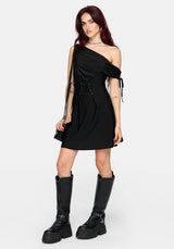 A woman modeling a black one-shoulder mini dress with a side tie detail, holding a black studded shoulder bag against a light gray background.