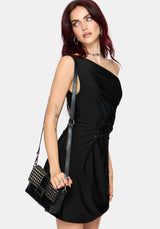 A woman modeling a black one-shoulder mini dress with a side tie detail, holding a black studded shoulder bag against a light gray background.