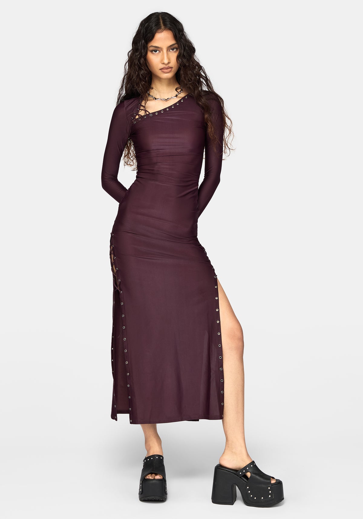 A woman modeling a long-sleeve, bodycon midi dress in deep plum purple with silver stud embellishments along the neckline, side seams, and hem, paired with black platform shoes against a light gray background.