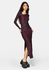 A woman modeling a long-sleeve, form-fitting midi dress in deep burgundy with a high slit, side lace-up details, and a wide black belt with silver buckles, paired with black platform studded sandals against a light gray studio background.