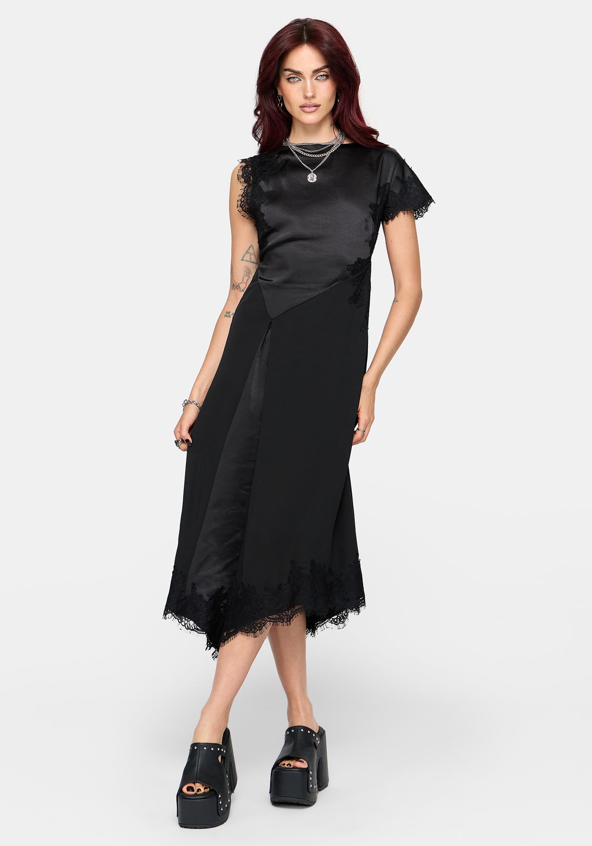 A model with red hair wearing a black satin midi dress with sheer ruffled short sleeves and a tattered hemline, paired with black platform shoes, against a light gray background.