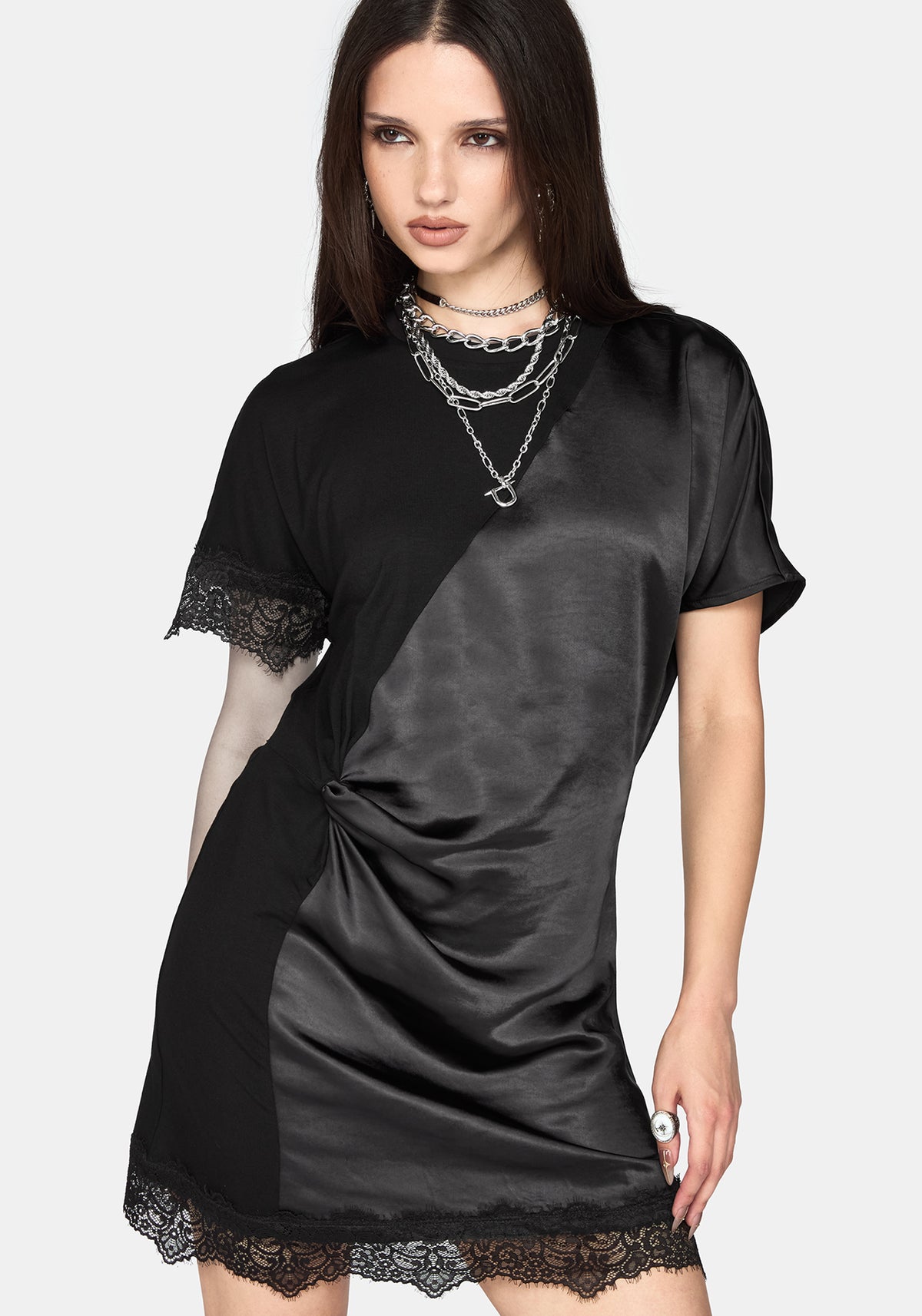 A woman modeling a black satin short-sleeve dress with lace trim on the sleeves and hem, styled with layered silver necklaces against a light gray background.