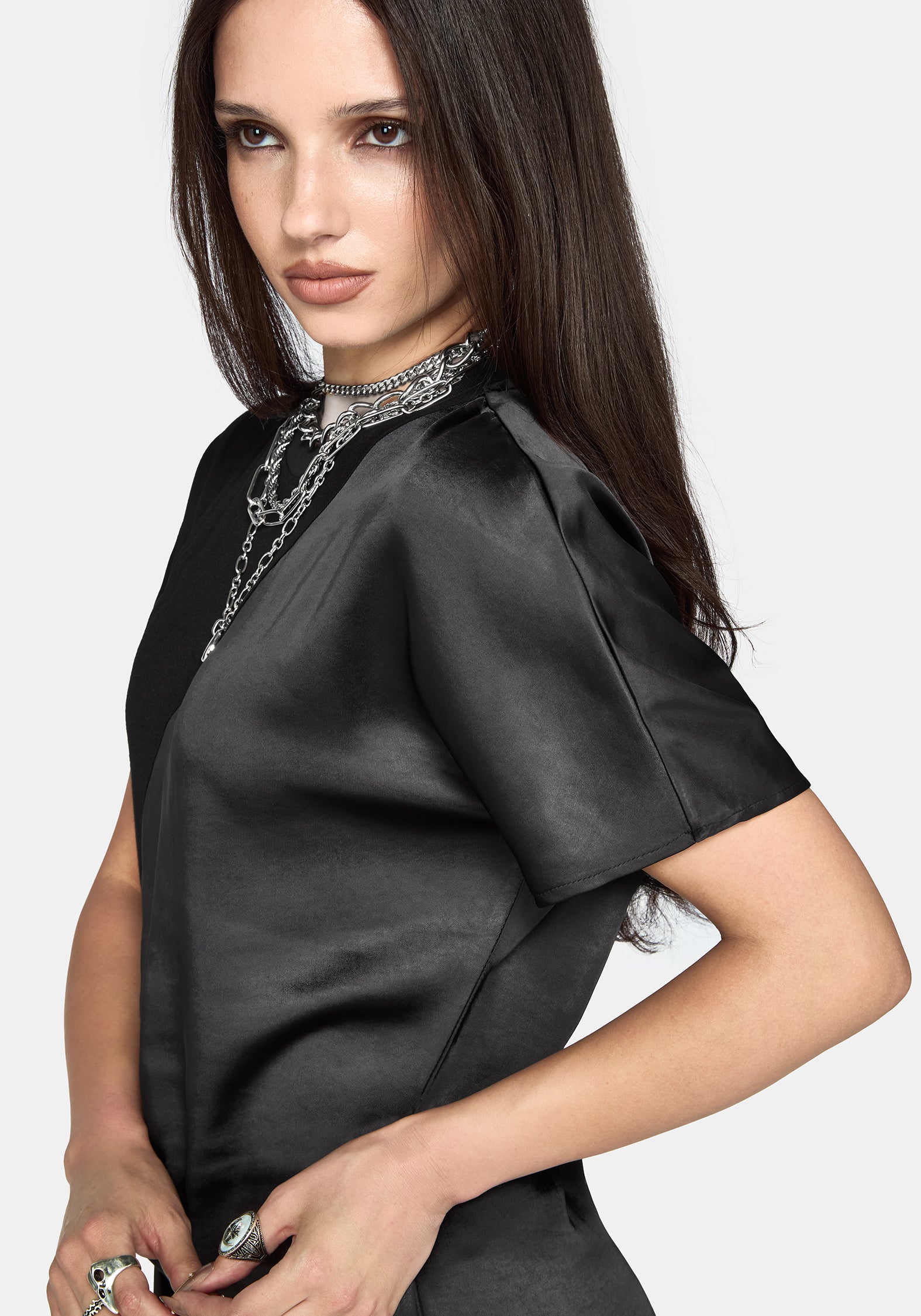 A woman modeling a black satin short-sleeve dress with lace trim on the sleeves and hem, styled with layered silver necklaces against a light gray background.