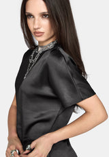 A woman modeling a black satin short-sleeve dress with lace trim on the sleeves and hem, styled with layered silver necklaces against a light gray background.