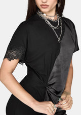 A woman modeling a black satin short-sleeve dress with lace trim on the sleeves and hem, styled with layered silver necklaces against a light gray background.