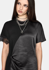 A woman wearing a black satin short-sleeve dress with lace trim on one sleeve and a twisted front detail, styled with layered silver chain necklaces against a light gray background.