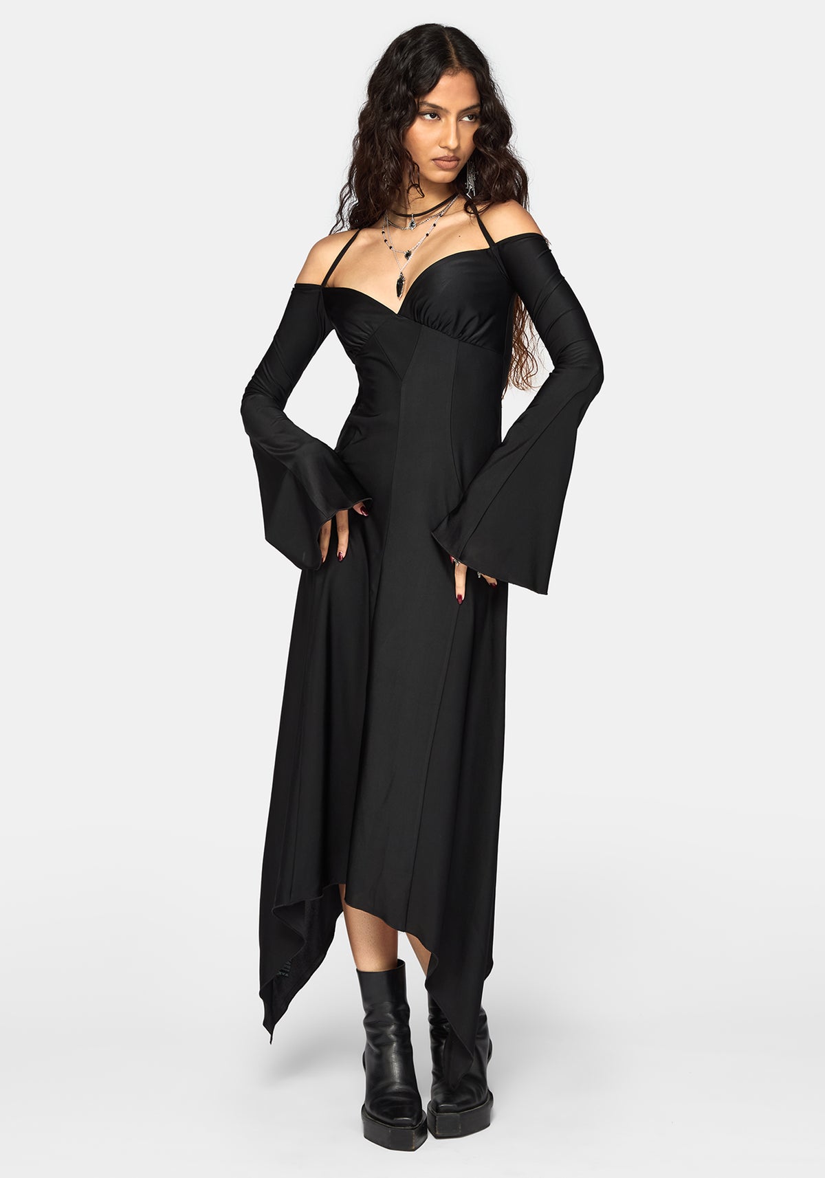 A woman modeling a black off-the-shoulder maxi dress with dramatic bell sleeves and an asymmetrical hem, paired with black platform ankle boots against a light gray studio background.