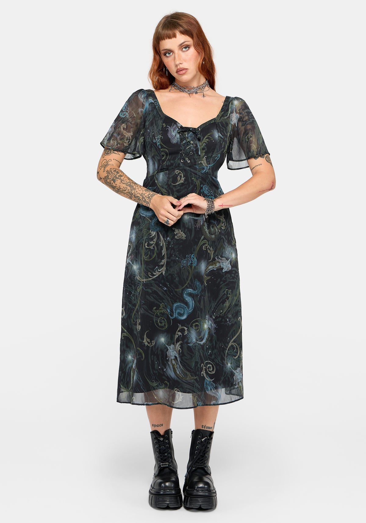 A model wearing a black midi-length sheer mesh dress with a blue and gold psychedelic swirl pattern, paired with black platform combat boots, against a light gray studio background.