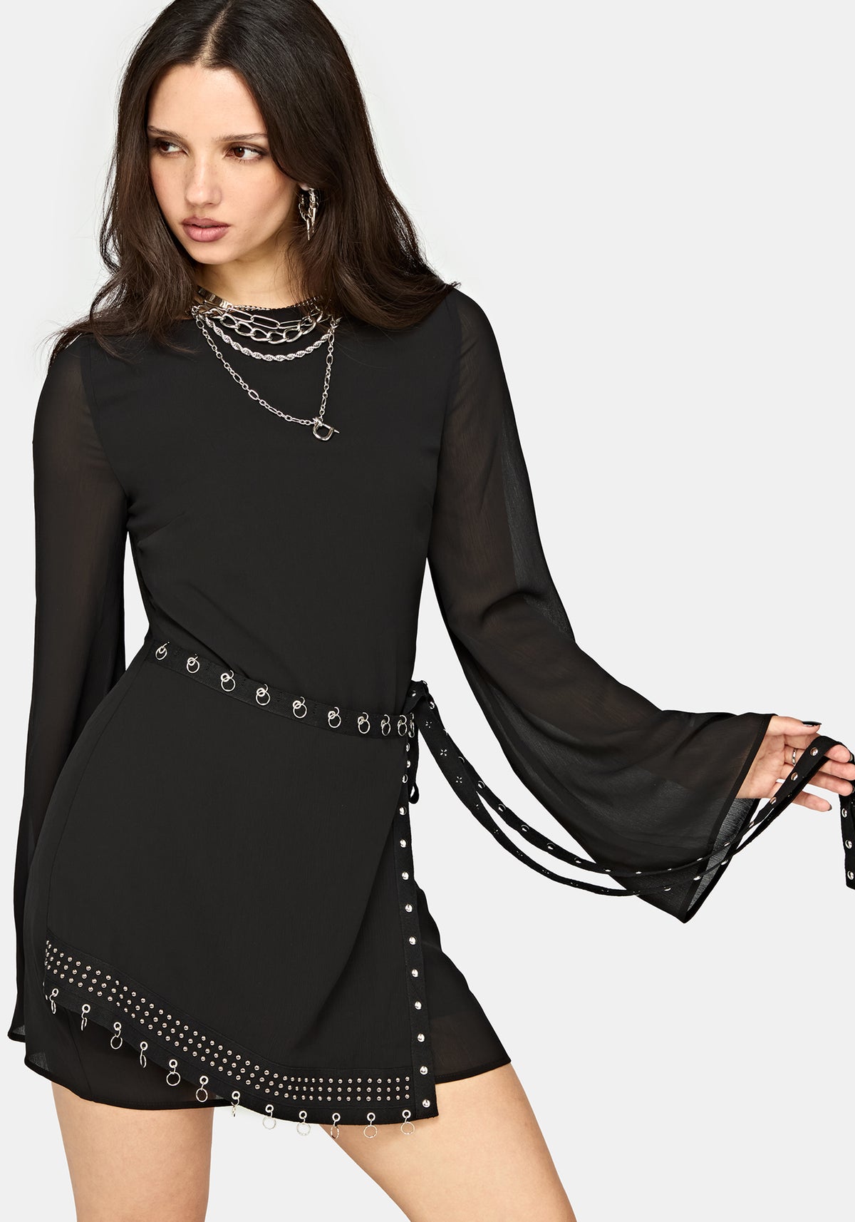 A woman modeling a black long-sleeve mini dress with sheer bell sleeves, a studded waist tie belt, and studded trim along the hem, against a light gray studio background.