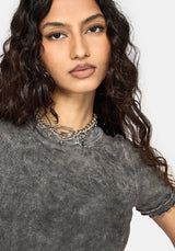 A close-up of a woman with long, wavy dark brown hair wearing a gray ribbed short-sleeve top and layered silver chain necklaces against a light gray background.