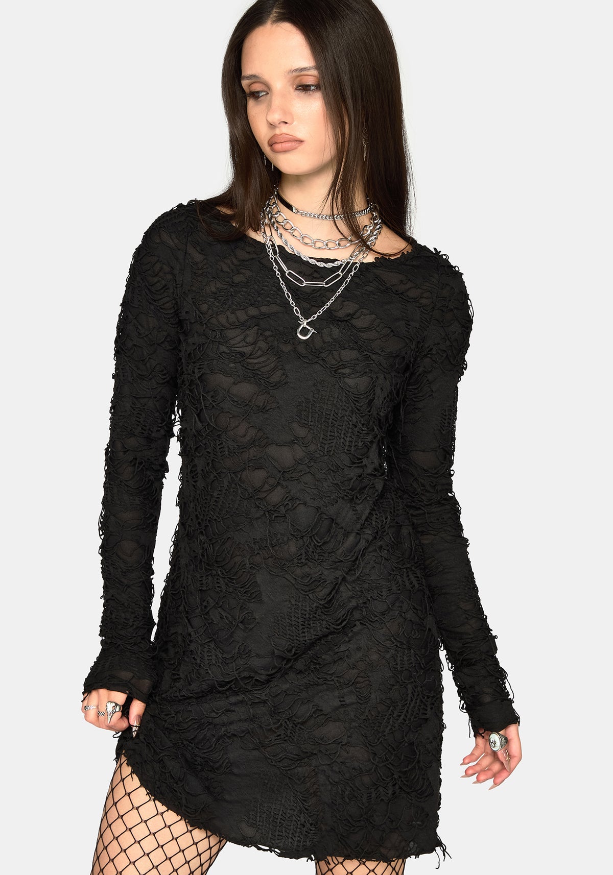 A woman modeling a black long-sleeve mini dress with frayed edges and layered silver necklaces, paired with black fishnet tights, against a light gray background.