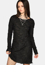 A woman modeling a black long-sleeve mini dress with frayed edges and layered silver necklaces, paired with black fishnet tights, against a light gray background.