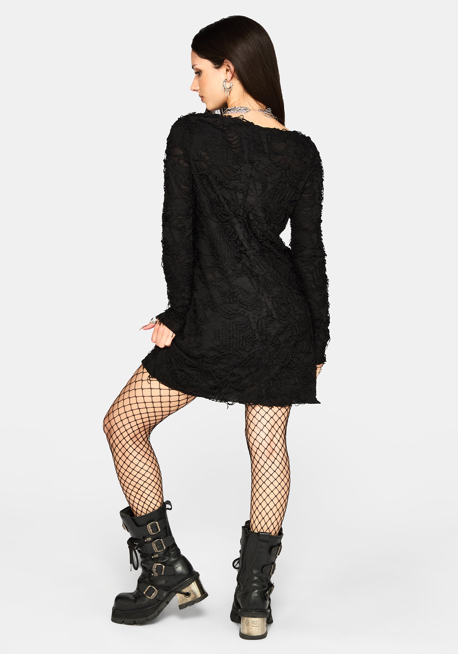 A woman posing from the back wearing a black textured long-sleeve mini dress, black fishnet stockings, and black platform lace-up boots with gold buckles, set against a plain light gray background.