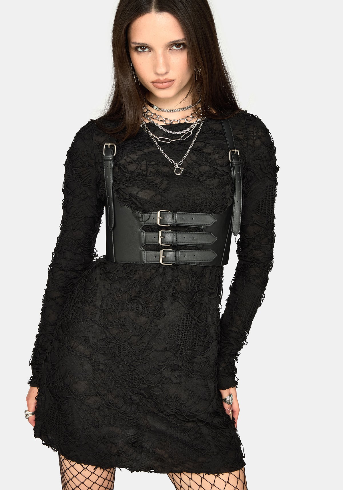 A model wearing a black long-sleeve mini dress with a wide black faux-leather belt featuring three silver buckles and matching shoulder straps, styled with layered silver necklaces and fishnet tights against a light gray background.