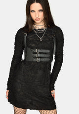 A model wearing a black long-sleeve mini dress with a wide black faux-leather belt featuring three silver buckles and matching shoulder straps, styled with layered silver necklaces and fishnet tights against a light gray background.