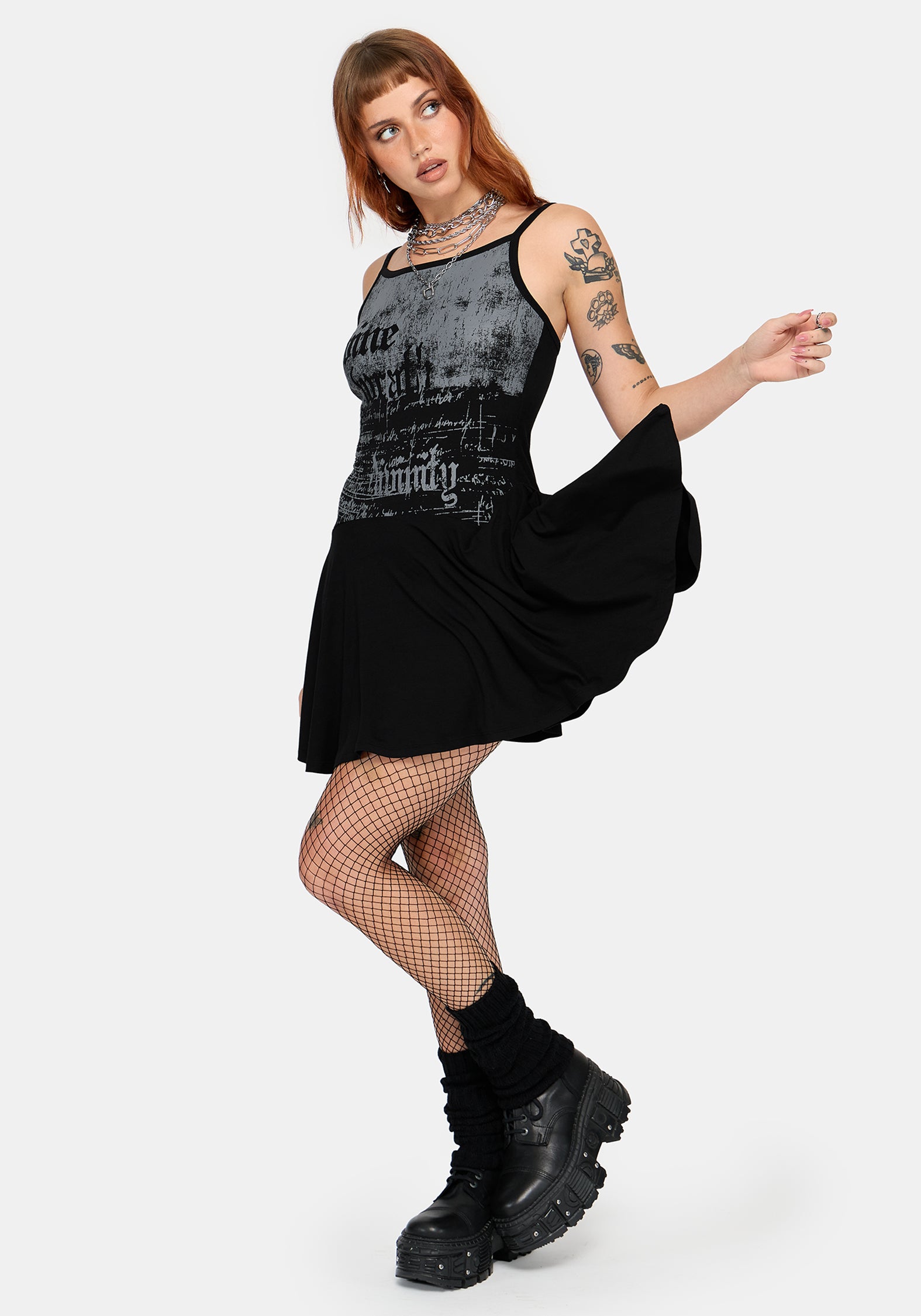 A model wearing a black sleeveless mini dress with a distressed gothic graphic print on the bodice, styled with fishnet tights, black socks, and chunky black boots against a light gray background.