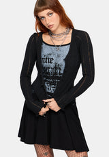 A model wearing a black long-sleeve ribbed cardigan over a black and gray graphic print mini dress and black, styled with layered silver necklaces and fishnet tights against a light gray background.
