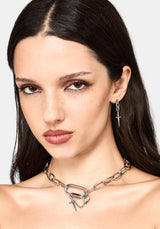 A close-up of a model wearing a silver chain necklace with a large open ring and dangling cross charms, along with a matching silver drop earring.