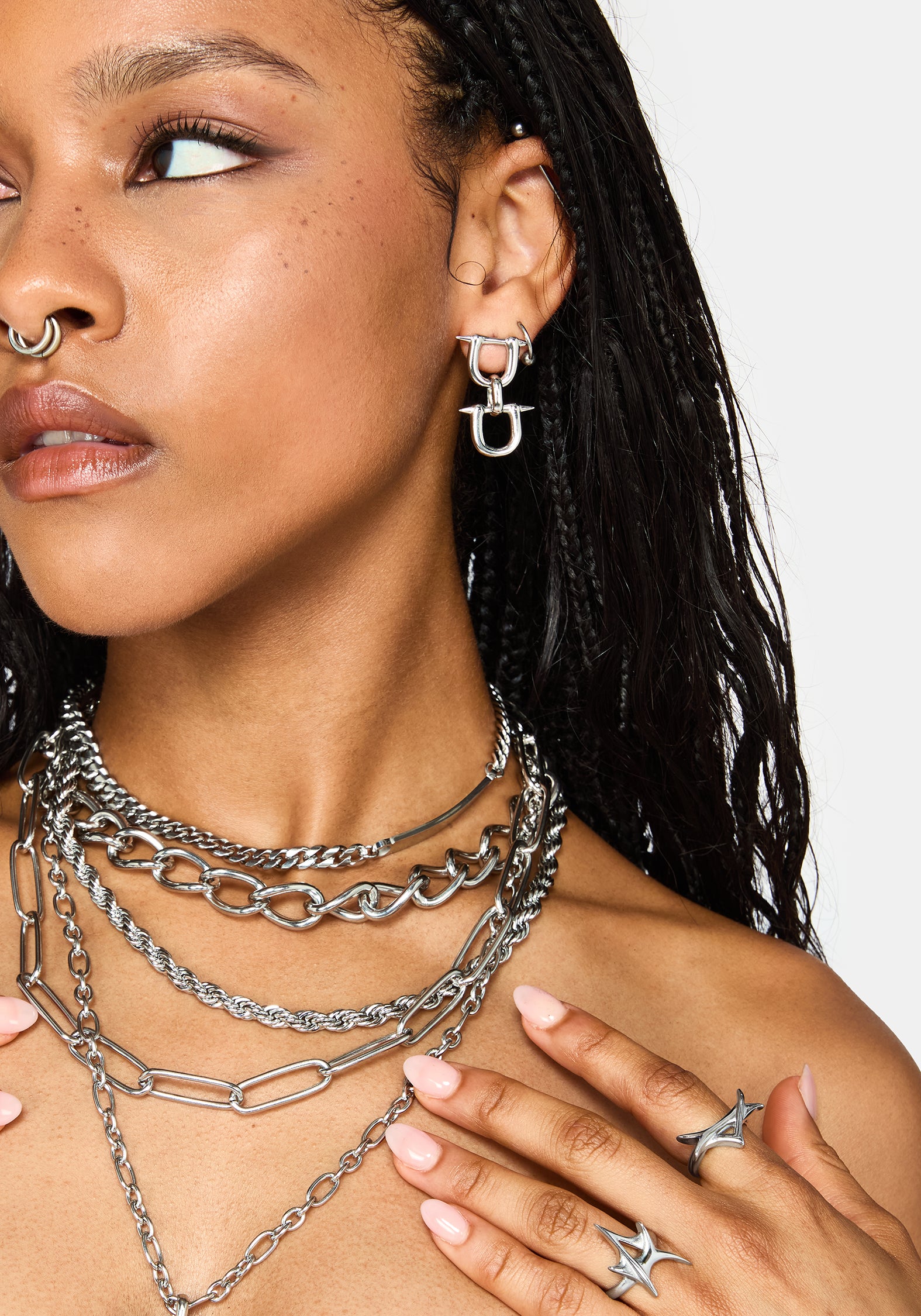 A close-up of a model wearing layered silver chain necklaces, a matching silver hoop earring with a stylized letter 'H' design, and silver star-shaped rings on her fingers.