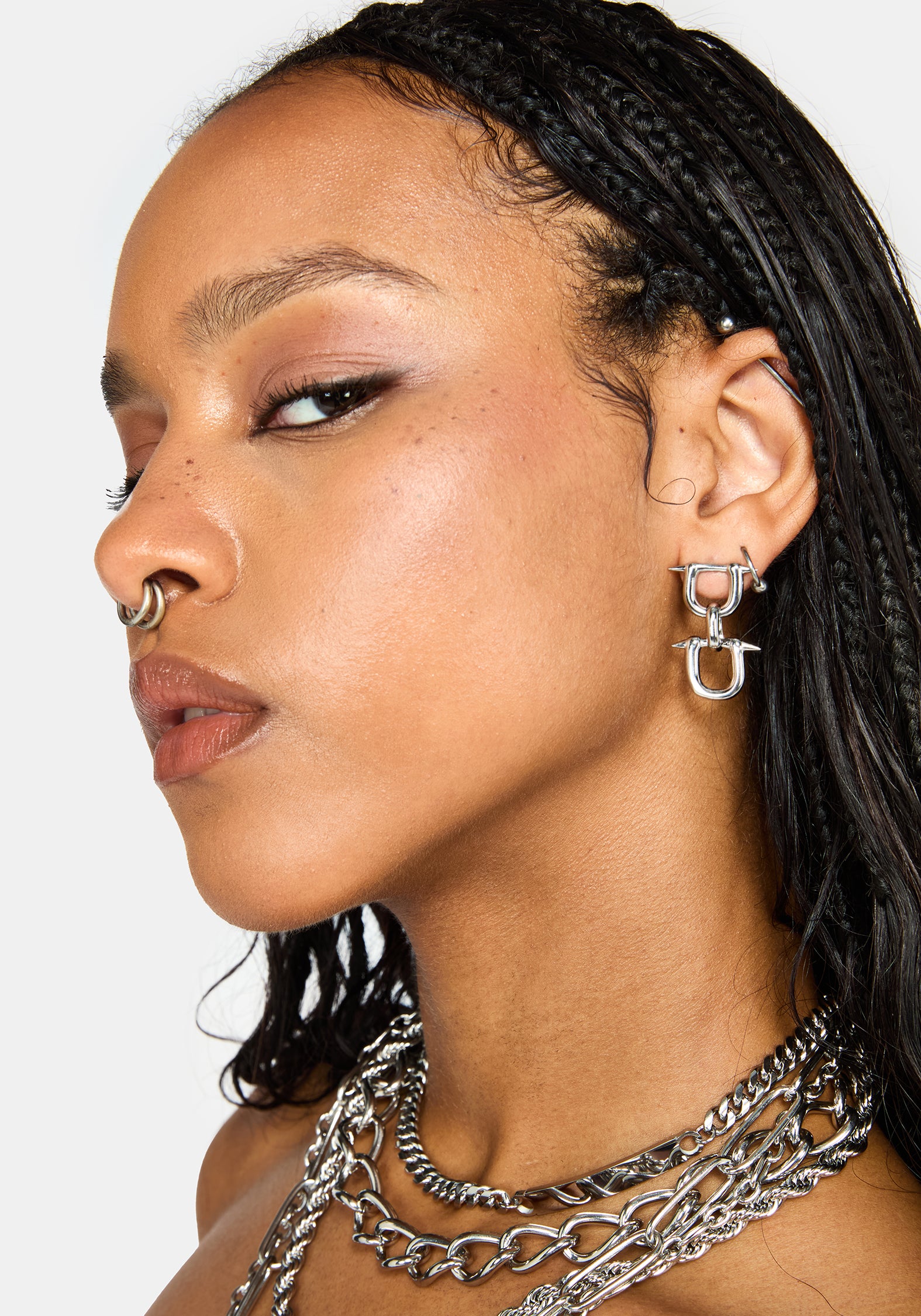 A close-up side profile of a model wearing a silver geometric hoop earring, a nose ring, and layered silver chain necklaces against a light gray background.