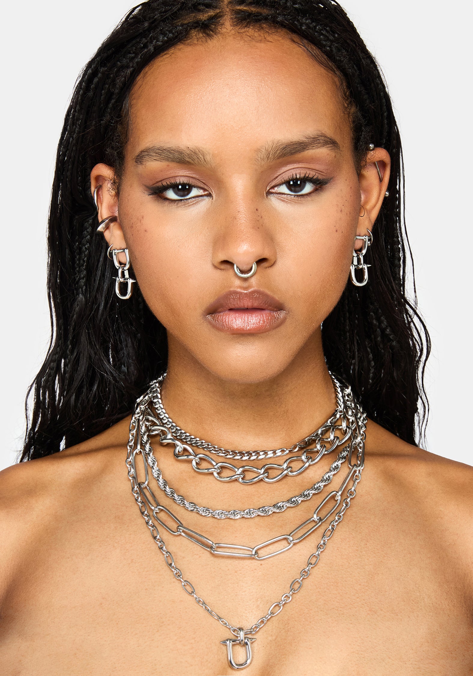 A close-up of a model wearing a layered stack of silver-tone chain necklaces, including chunky cable, paperclip, and delicate link styles, along with matching silver hoop earrings.