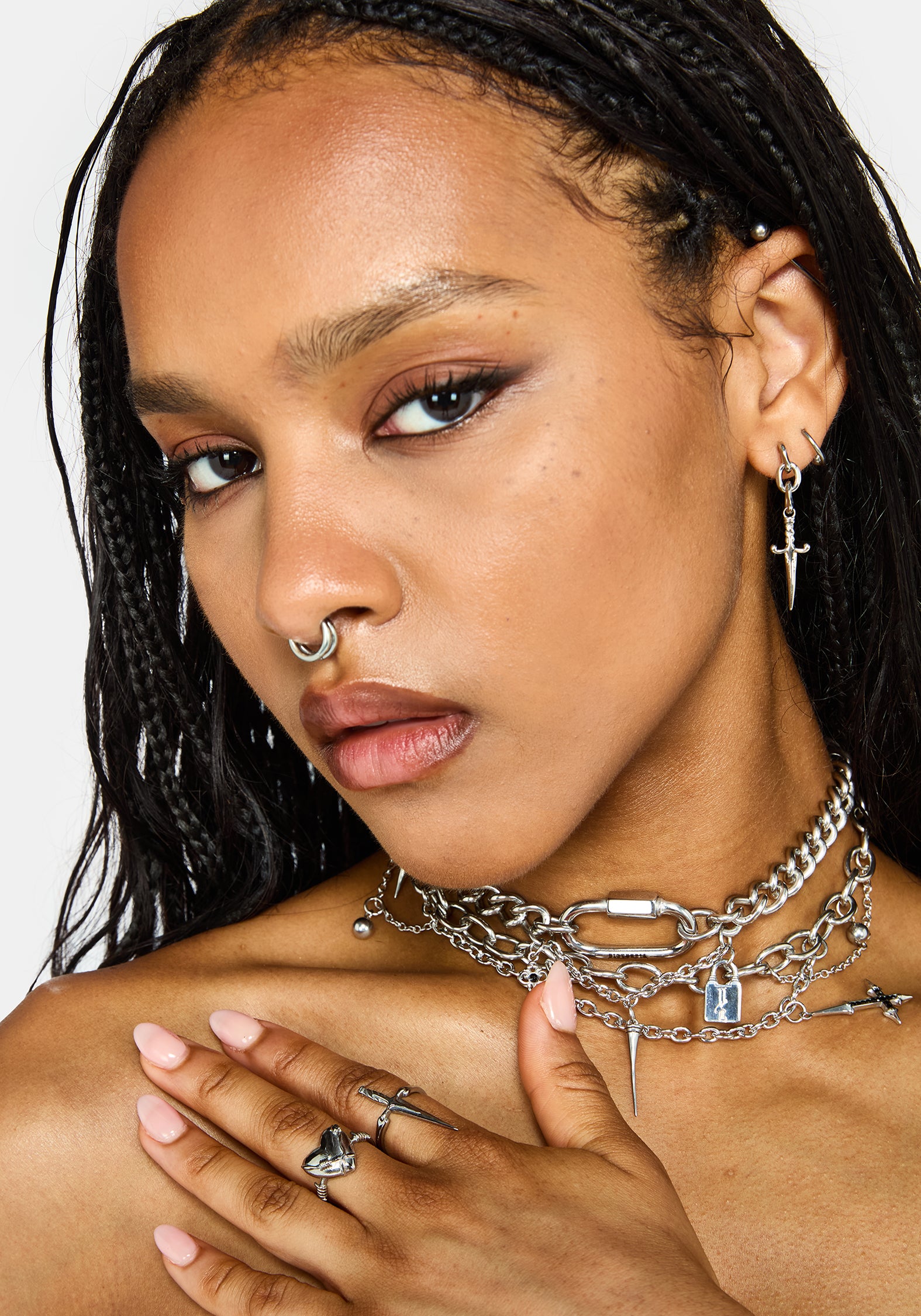 A close-up portrait of a model wearing multiple silver-toned piercings and jewelry, including a chain choker with padlock and star charms, a hoop nose ring, a nose lobe piercing, and cross earrings.