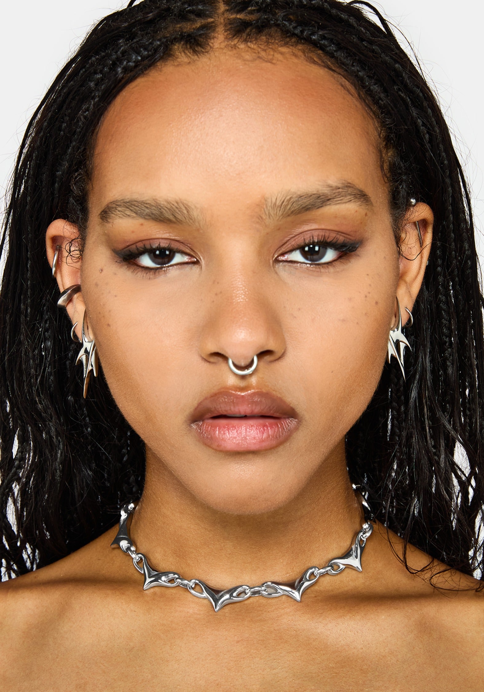 A close-up portrait of a model wearing a silver spike choker necklace, matching silver hoop nose ring, and silver star-shaped earrings against a plain light gray background.