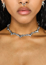 A close-up of a model wearing a silver gothic-style chain choker necklace with sharp, pointed bat-wing-like links.