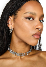 A close-up of a model wearing a chunky silver chain choker, along with matching silver hoop earrings and a nose ring, against a plain white background.
