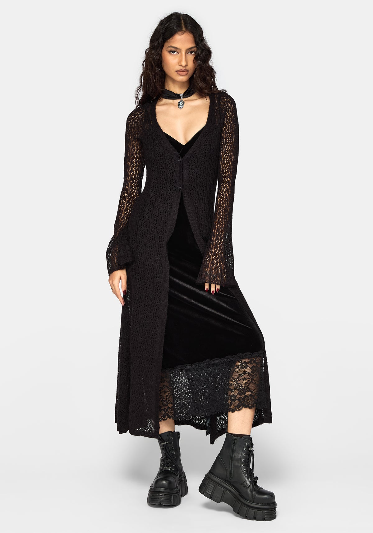 A model wearing a black long-sleeve lace-trimmed cardigan over a black velvet midi dress, styled with black combat boots and a choker, against a light gray studio background.