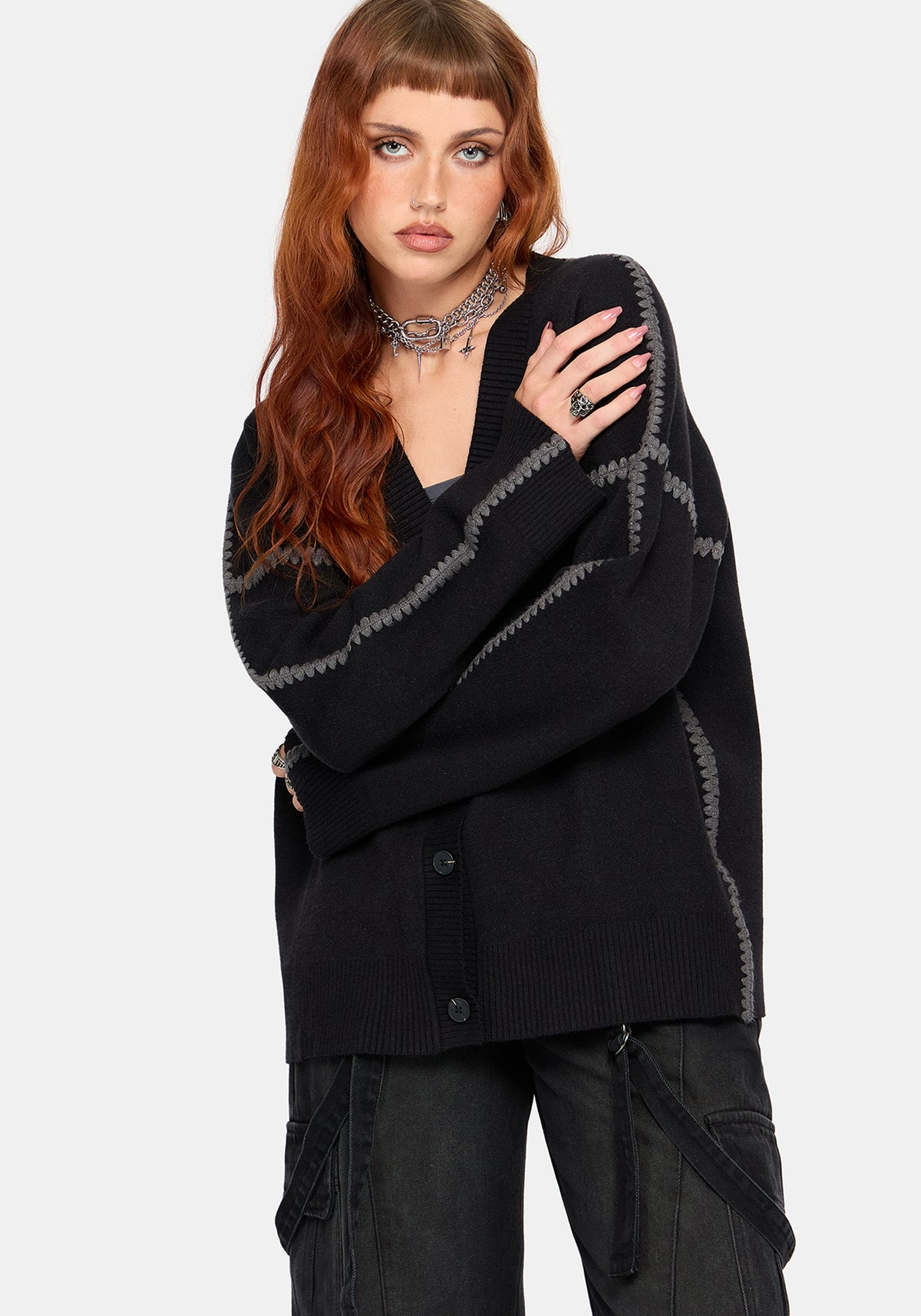 A woman modeling a black knit cardigan with gray geometric patterned details on the sleeves and sides, paired with dark cargo pants, against a light gray studio background.